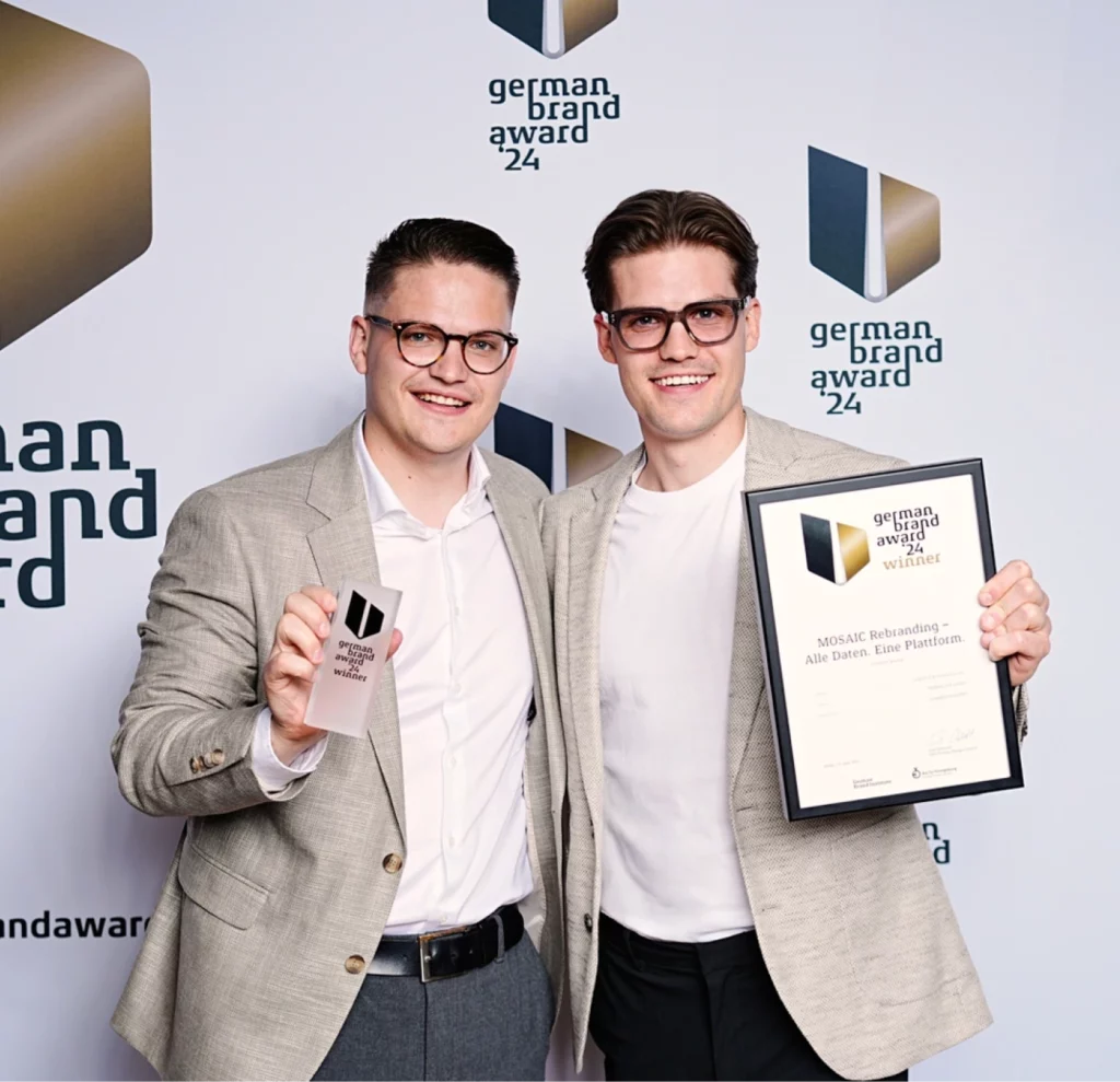 Zwei Männer mit Brillen und hellen Jacken halten eine Auszeichnung und eine Urkunde beim German Brand Award 2024