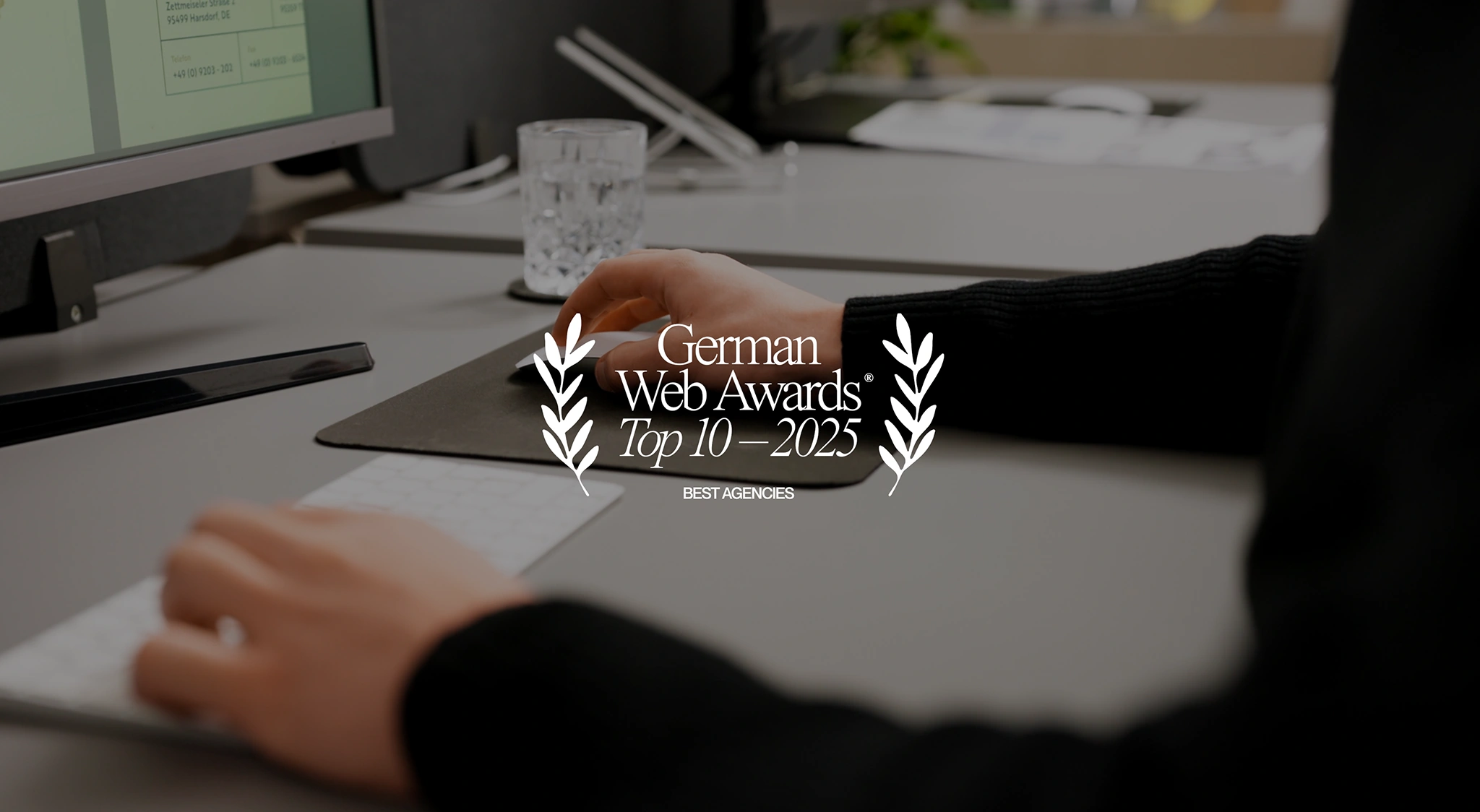 Eine Hand tippt auf einer Tastatur, während die andere Hand auf einem Mauspad liegt. Im Vordergrund steht der Text 'German Web Awards Top 10 - 2025' umrahmt von Lorbeerzweigen.